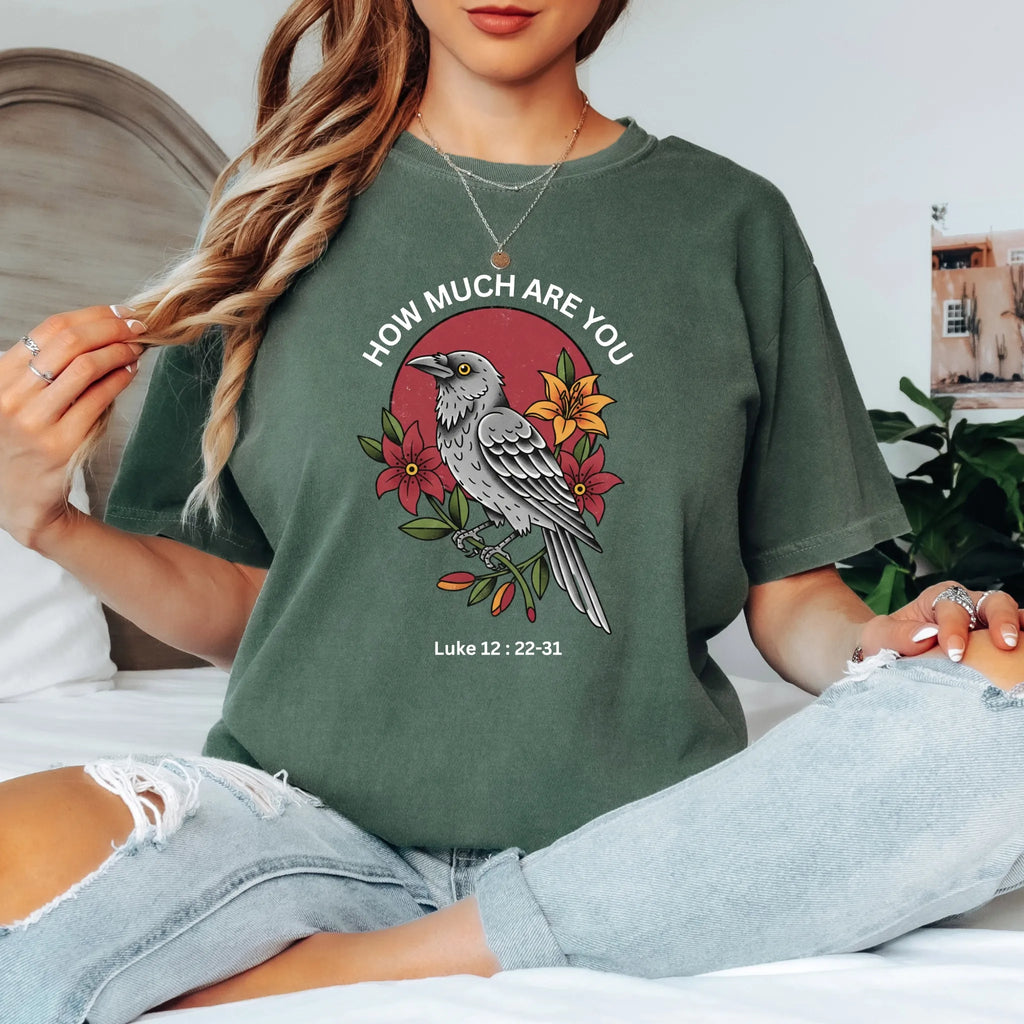 A woman sitting on a bed wearing a moss colour graphic t-shirt with a bird and floral design, paired with light-wash distressed jeans.