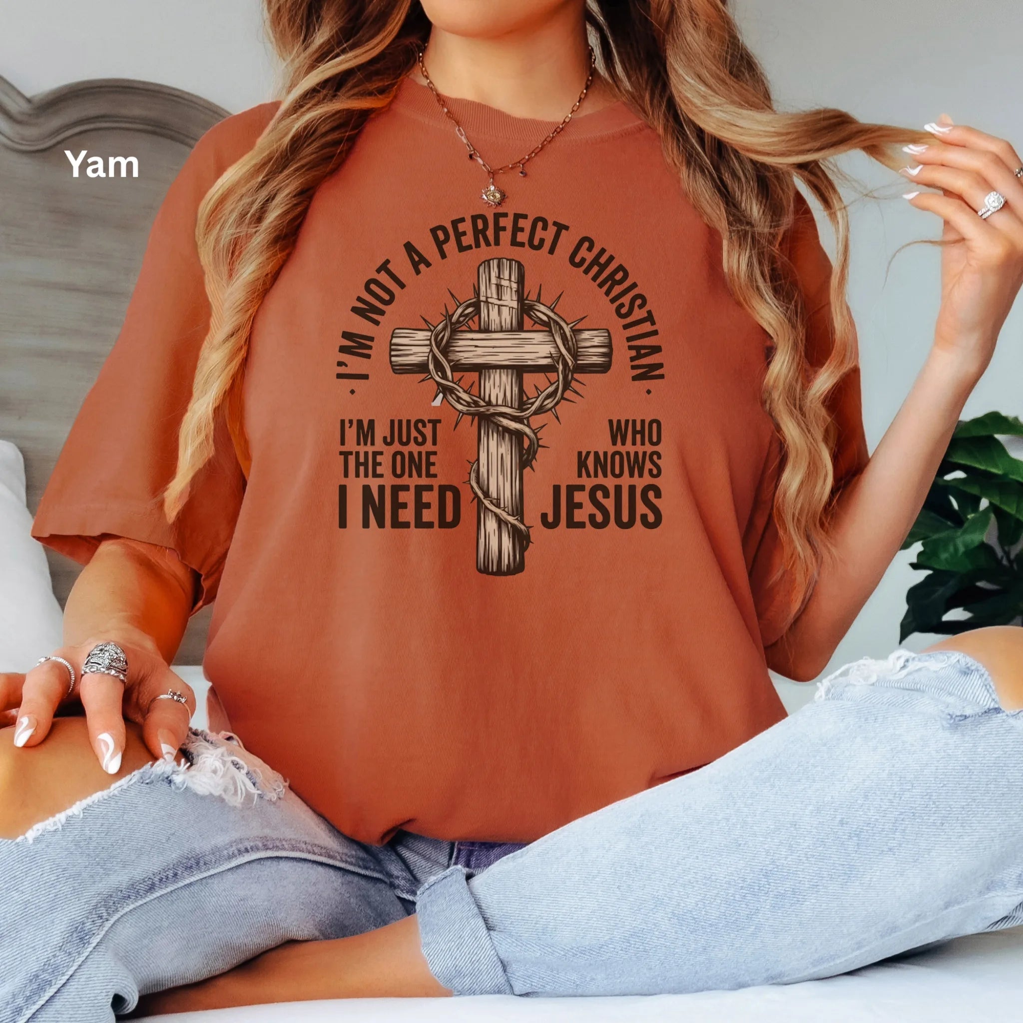 A woman wearing a yam colour graphic t-shirt with a cross and the text 'I'M NOT A PERFECT CHRISTIAN I'M JUST THE ONE I NEED WHO KNOWS JESUS'. She is wearing light-wash ripped jeans and sitting on a bed.